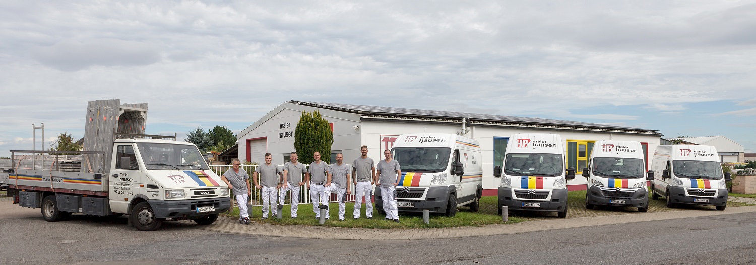 Malerfahrzeuge vor einem Betriebsgebäude mit Firmenlogo "Maler Hauser". Es sind verschiedene Fahrzeuge zu sehen, darunter ein Lastwagen mit einer Ladefläche und mehrere Kleintransporter. In der Mitte steht das Team der Firma versammelt. Das Betriebsgelände hat einen asphaltierten Parkplatz und Grünflächen. Das Gebäude im Hintergrund ist weiß mit roten Akzenten und einem großen Fensterbereich.