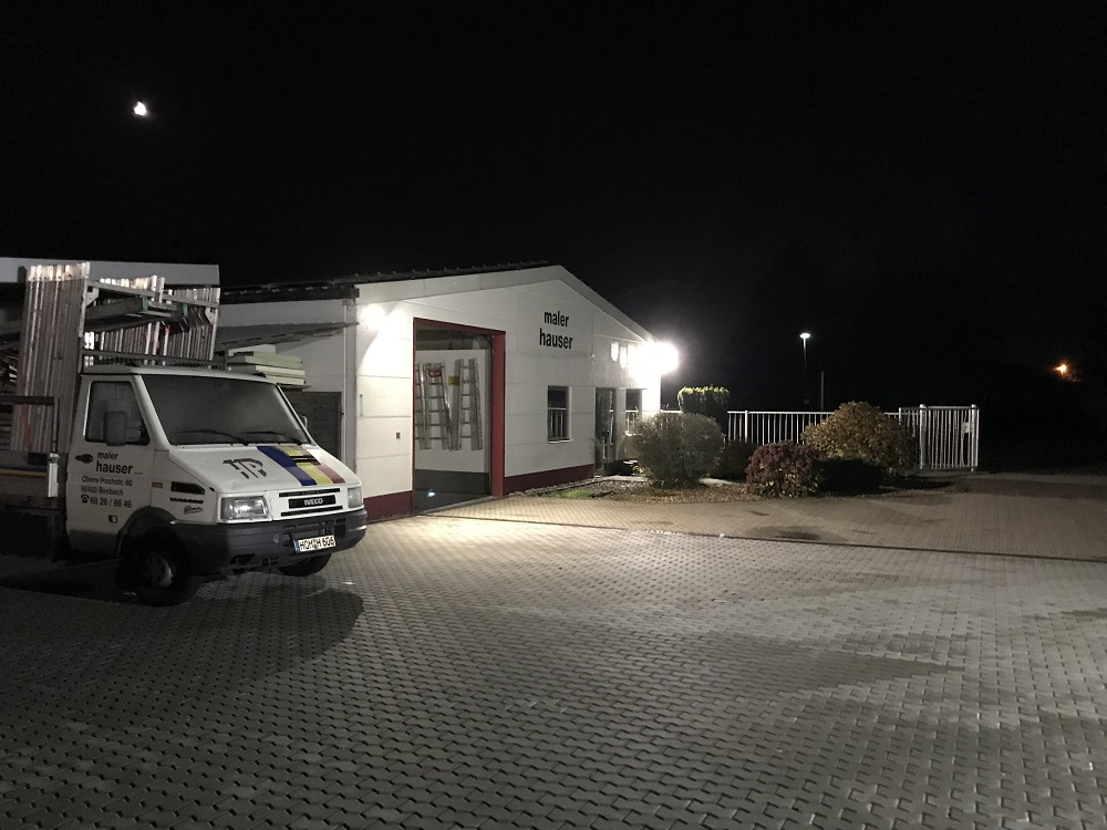 Nächtliche Ansicht eines Betriebsgebäudes mit der Aufschrift "Maler Hauser". Vor dem Gebäude steht ein Malerfahrzeug mit Ladefläche. Das Gelände ist gepflastert und durch Straßenlaternen beleuchtet. Das Betriebsgebäude hat eine weiße Fassade mit roten Akzenten und eine Glastür. Der Himmel zeigt einen sichtbaren Mond.