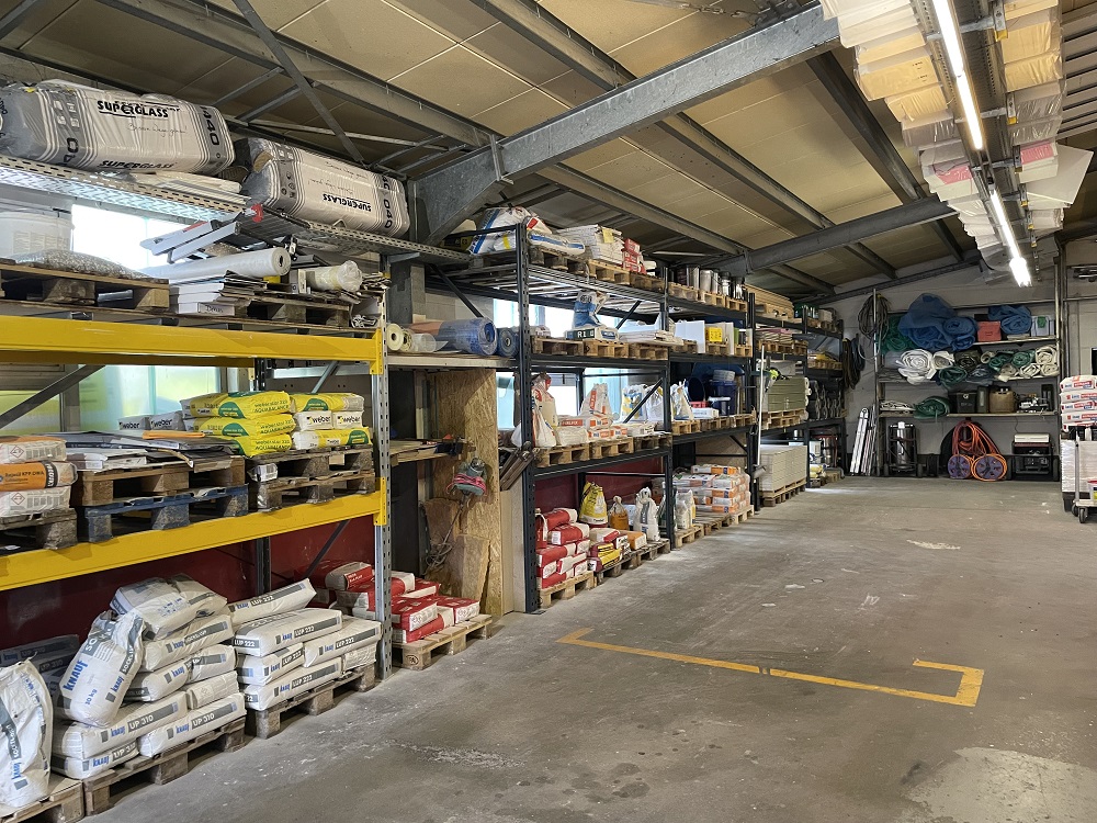 Innenansicht einer Lagerhalle mit mehreren Regalen, die mit Baumaterialien bestückt sind. Es gibt Säcke mit Beschriftungen, Eimer, Werkzeuge und diverse andere Bauartikel. Die Regale sind in verschiedenen Farben, darunter Gelb und Blau. Der Boden der Halle ist betoniert und mit gelben Markierungen versehen. Das Dach besteht aus Metallträgern, und es gibt mehrere Beleuchtungskörper, die die Halle erhellen.