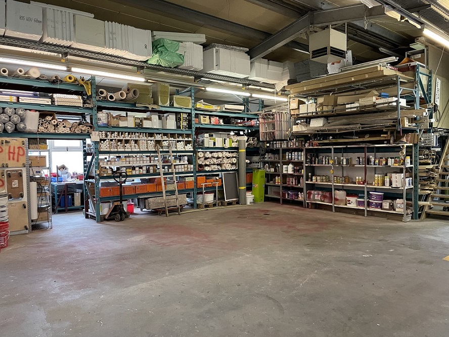 Innenansicht einer Lagerhalle mit mehreren Regalen, die mit Baumaterialien bestückt sind. Es gibt Säcke mit Beschriftungen, Eimer, Werkzeuge und diverse andere Bauartikel. Die Regale sind in verschiedenen Farben, darunter Gelb und Blau. Der Boden der Halle ist betoniert und mit gelben Markierungen versehen. Das Dach besteht aus Metallträgern, und es gibt mehrere Beleuchtungskörper, die die Halle erhellen.