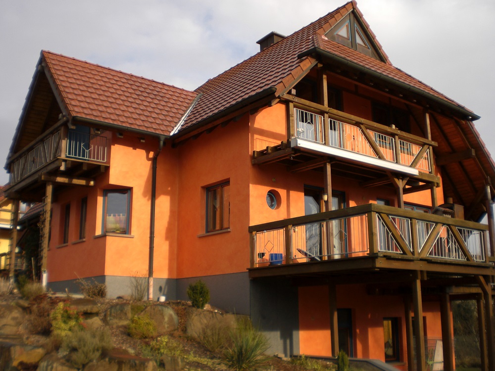 Ein charmantes zweistöckiges Haus mit traditionellem europäischem Design. Das Haus hat eine auffällige orangefarbene Fassade, die von Holzbalkonen und -verkleidungen akzentuiert wird. Das steil geneigte Dach, das typisch für viele europäische Länderhäuser ist, ist mit roten Ziegeln gedeckt. Es gibt mehrere Balkone, die einen schönen Blick auf die Umgebung bieten könnten. Das Haus scheint in einer hügeligen Gegend zu stehen, und der Himmel darüber ist teilweise bewölkt, was dem Bild eine gemütliche und warme Atmosphäre verleiht. Es wirkt wie ein perfekter Rückzugsort zum Entspannen und Genießen der Natur.