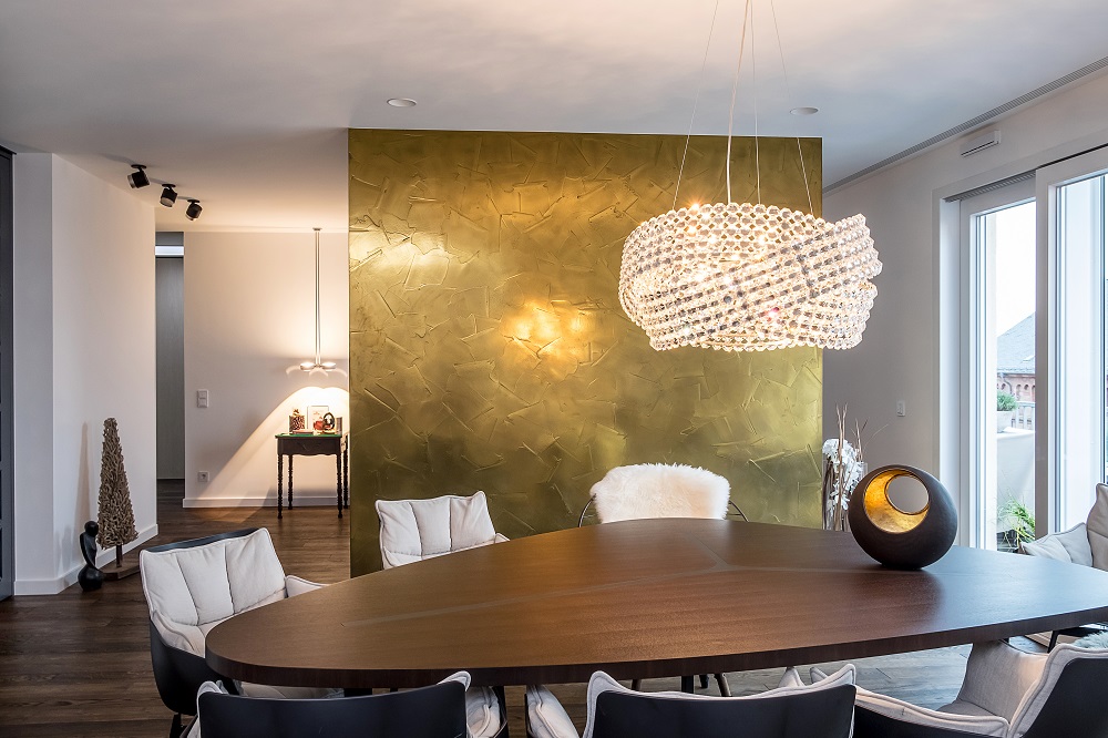 Ein stilvoll eingerichteter Essbereich mit einem großen dunklen Holztisch und mehreren weißen Stühlen. Über dem Tisch hängt eine beeindruckende Pendelleuchte mit zahlreichen Kristallen. Hinter dem Tisch steht eine große, goldene Wand mit einer rauen, texturierten Oberfläche, die durch zwei Wandlampen beleuchtet wird. Im Hintergrund befindet sich ein kleiner Tisch mit Dekorationsartikeln und eine Glasfront bietet einen Blick nach draußen. Der gesamte Raum strahlt eine Atmosphäre von Luxus und Eleganz aus.