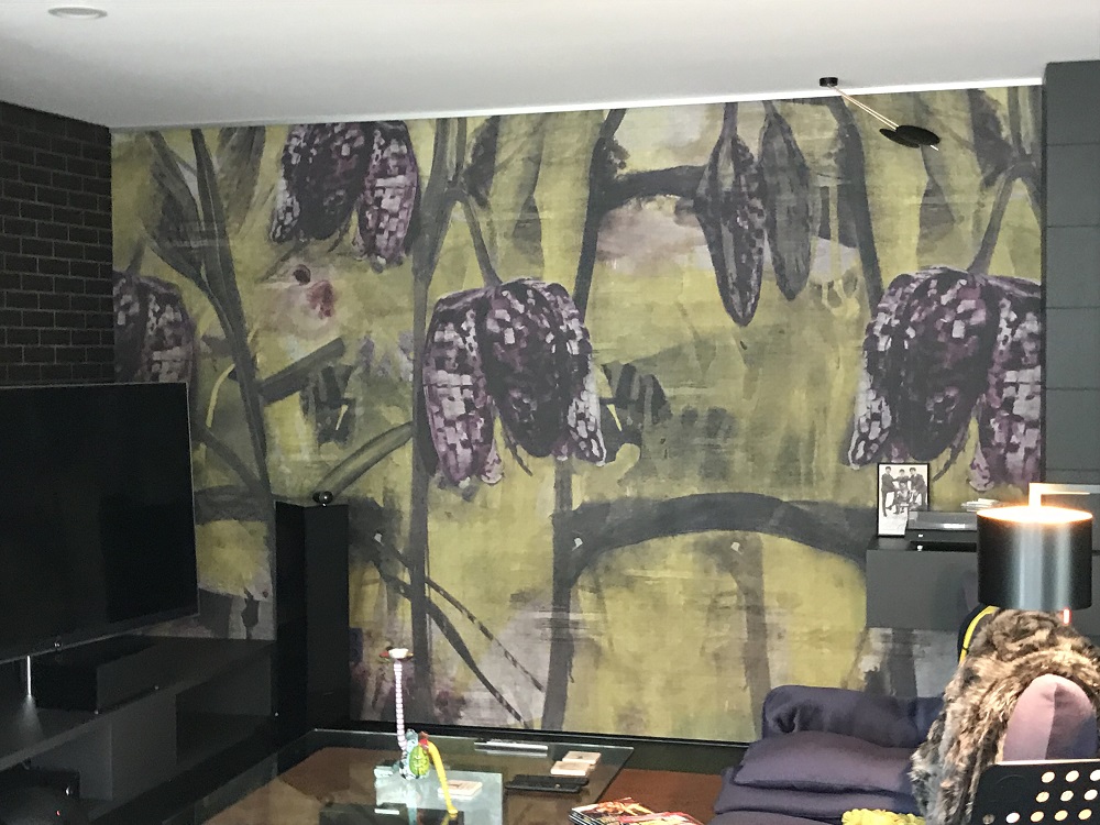 Ein modernes Wohnzimmer mit einem markanten Wandbild, das grüne Pflanzen und dunkelviolette Zapfen zeigt. Vor der Wand steht ein schwarzes Unterhaltungssystem mit einem Fernseher. Daneben befindet sich eine gemütliche Sitzecke mit einem blauen Sessel und einem Glas-Couchtisch, auf dem verschiedene Gegenstände liegen. Die Zimmeratmosphäre wird durch eine stehende Lampe und die dunklen Wände betont. Der Raum kombiniert moderne Einrichtungselemente mit dem auffälligen Naturmotiv der Wand.