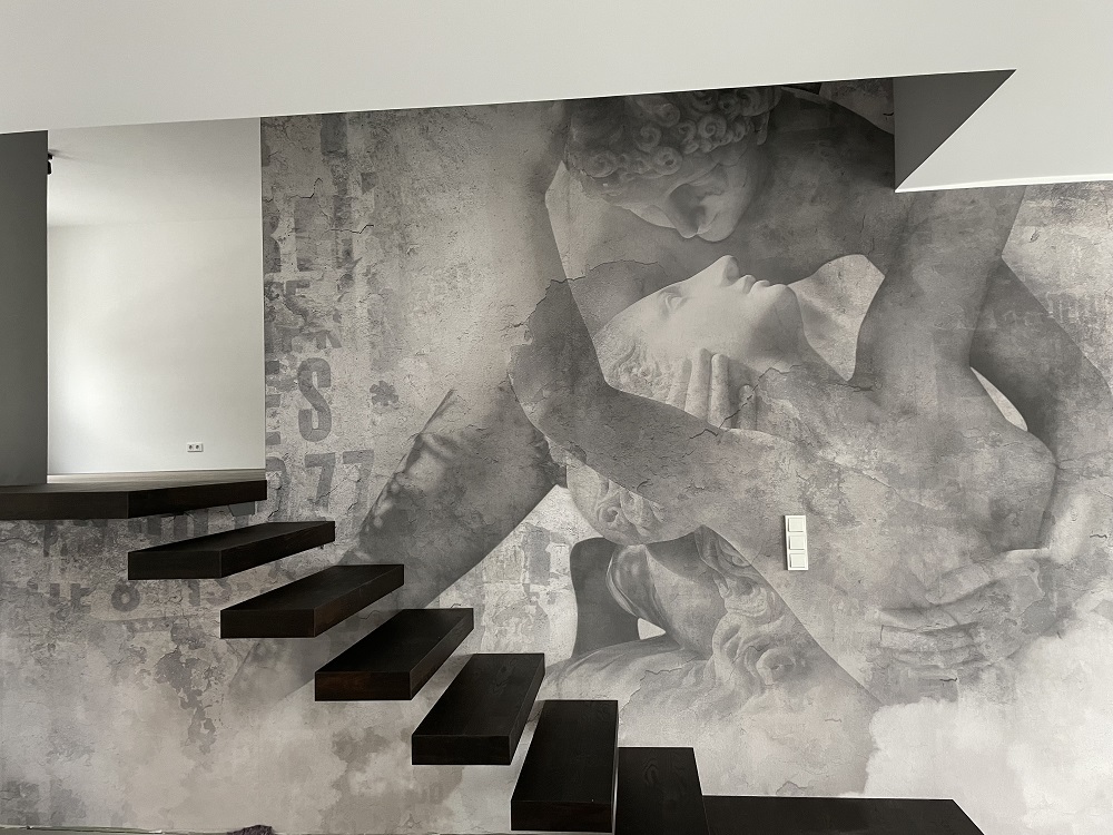 Wanddekoration mit schwarz-weißem Druck einer klassischen Statue und sichtbaren Buchstaben neben einer modernen schwarzen Treppe.