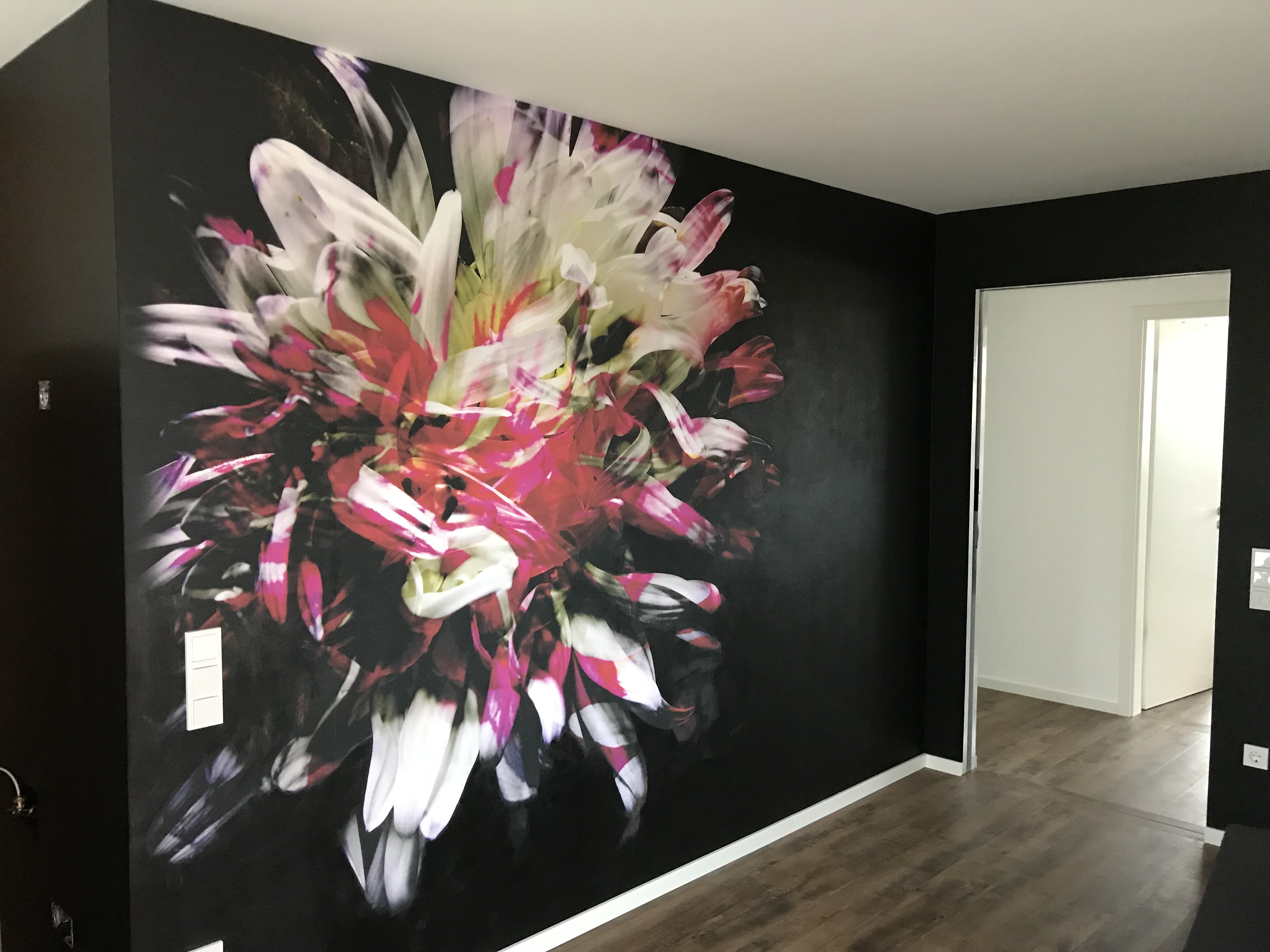 Schwarze Wand mit großem bunten Blumen-Gemälde, Holzboden und Tür im Hintergrund.