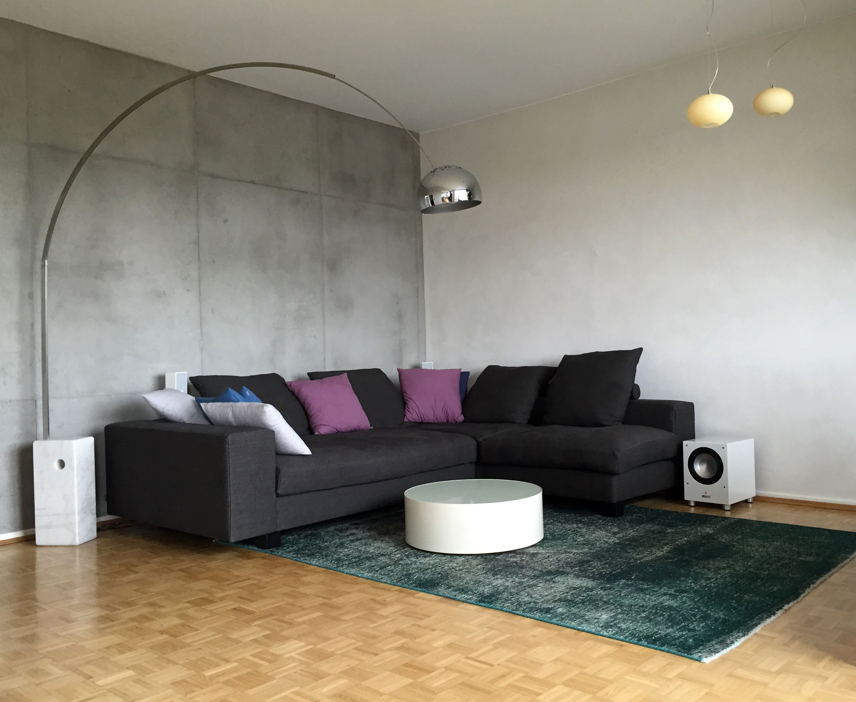 Wohnzimmer mit dunkelgrauem Sofa, bunten Kissen, weißem runden Couchtisch auf grünem Teppich, Betonwand-Optik und Bogenlampe.