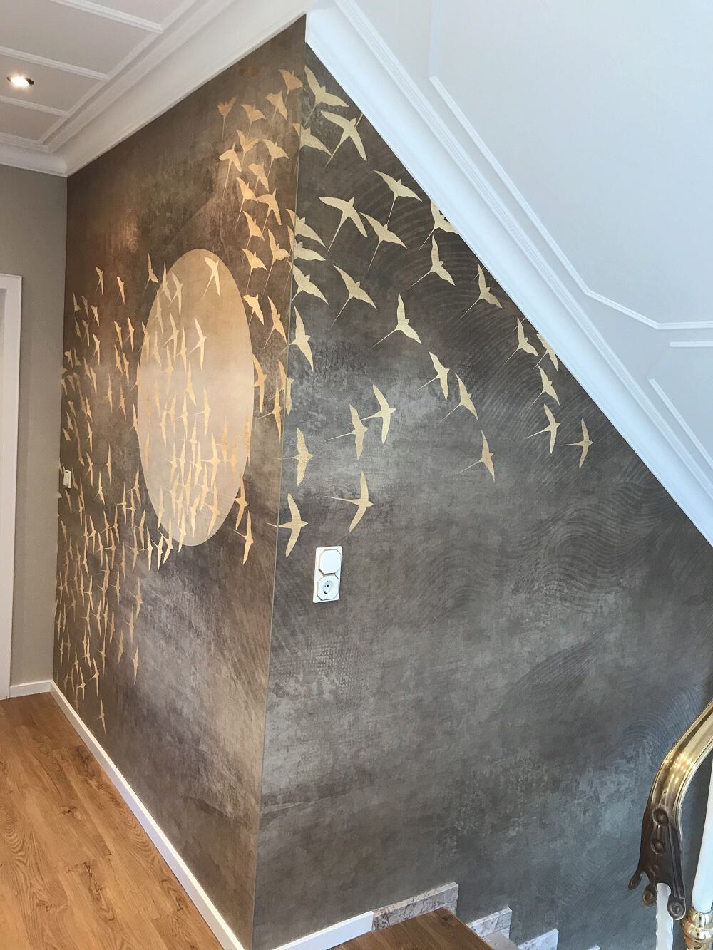 Ein modernes Wohnzimmer mit einem markanten Wandbild, das grüne Pflanzen und dunkelviolette Zapfen zeigt. Vor der Wand steht ein schwarzes Unterhaltungssystem mit einem Fernseher. Daneben befindet sich eine gemütliche Sitzecke mit einem blauen Sessel und einem Glas-Couchtisch, auf dem verschiedene Gegenstände liegen. Die Zimmeratmosphäre wird durch eine stehende Lampe und die dunklen Wände betont. Der Raum kombiniert moderne Einrichtungselemente mit dem auffälligen Naturmotiv der Wand.